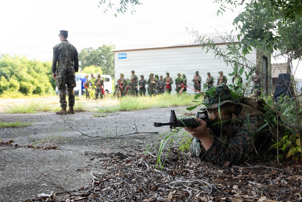 A military training exercise is underway at what appears to be a simulated urban environment. A soldier in camouflage is in a prone position aiming a rifle while concealed by vegetation near a building corner. In the background, a group of uniformed personnel observe the exercise while an instructor or evaluator stands nearby.