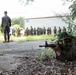 A military training exercise is underway at what appears to be a simulated urban environment. A soldier in camouflage is in a prone position aiming a rifle while concealed by vegetation near a building corner. In the background, a group of uniformed personnel observe the exercise while an instructor or evaluator stands nearby.