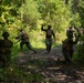 A group of military personnel in woodland camouflage uniforms are conducting a field training exercise along a dirt trail in a dense forested area. Several individuals are moving tactically, with at least one person giving hand signals, suggesting a patrol or movement-to-contact drill. The setting appears to be a training environment during daylight hours in a temperate forest.