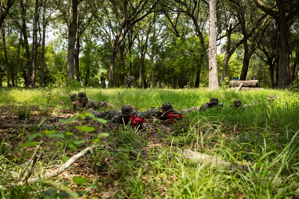 Military personnel in camouflage uniforms are conducting a training exercise in a wooded, grassy area. Several soldiers are in the prone position in the foreground holding red training weapons (MILES gear or dummy rifles), while two additional personnel stand in the background observing or supervising. The red-colored weapons indicate this is a training or simulation exercise rather than a live combat scenario.