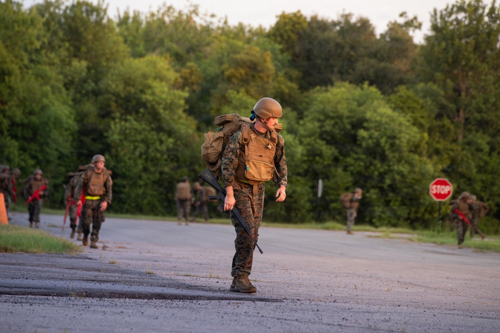 A group of military personnel in full combat gear are conducting a road march along a paved road bordered by dense green trees. The lead soldier is prominently visible in the foreground, carrying a rifle and wearing a tactical vest, helmet, and backpack. Additional soldiers are visible in the background, similarly equipped, suggesting a training exercise or field operation.