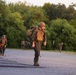 A group of military personnel in full combat gear are conducting a road march along a paved road bordered by dense green trees. The lead soldier is prominently visible in the foreground, carrying a rifle and wearing a tactical vest, helmet, and backpack. Additional soldiers are visible in the background, similarly equipped, suggesting a training exercise or field operation.