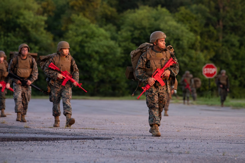A group of military personnel in camouflage uniforms are conducting a training exercise, marching along a paved road while carrying bright red training rifles. The red-colored weapons indicate these are inert training replicas, not live firearms, consistent with military recruit or unit training scenarios. The setting appears to be a wooded training facility during early morning or evening light.