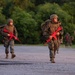 A group of military personnel in camouflage uniforms are conducting a training exercise, marching along a paved road while carrying bright red training rifles. The red-colored weapons indicate these are inert training replicas, not live firearms, consistent with military recruit or unit training scenarios. The setting appears to be a wooded training facility during early morning or evening light.