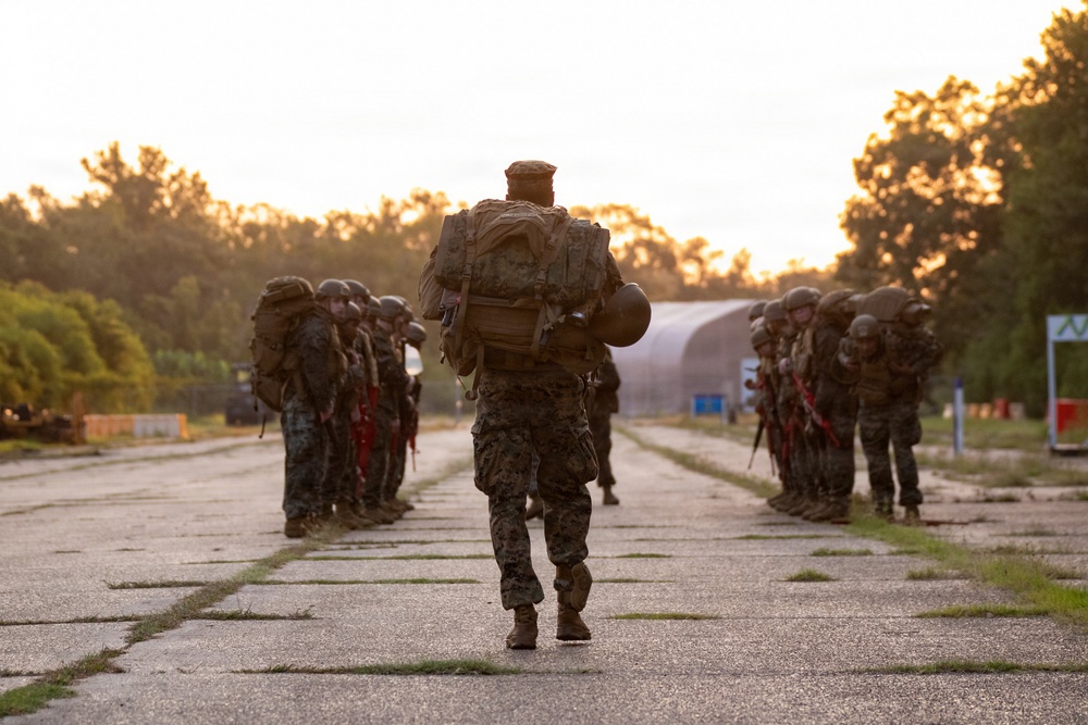 A military formation is assembled along both sides of a concrete road or airstrip during golden hour lighting conditions. A single service member carrying a large tactical backpack and helmet walks down the center of the formation, appearing to be an instructor or leader conducting an inspection or movement drill. The personnel are wearing camouflage uniforms consistent with U.S. Marine Corps MARPAT pattern and are equipped with rifles featuring red training barrel indicators.