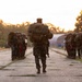 A military formation is assembled along both sides of a concrete road or airstrip during golden hour lighting conditions. A single service member carrying a large tactical backpack and helmet walks down the center of the formation, appearing to be an instructor or leader conducting an inspection or movement drill. The personnel are wearing camouflage uniforms consistent with U.S. Marine Corps MARPAT pattern and are equipped with rifles featuring red training barrel indicators.