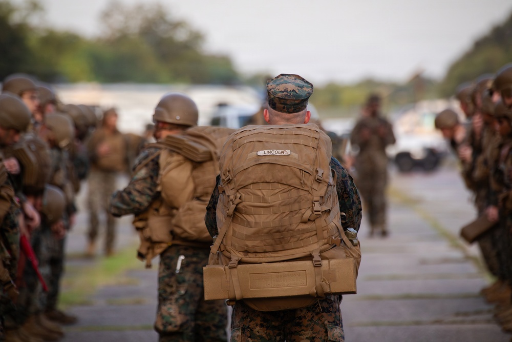 The image shows a group of military personnel, likely U.S. Marines based on their woodland MARPAT camouflage uniforms, assembled on a paved area such as a tarmac or road. The central figure, whose backpack bears the name tape 'LINCOLN,' is wearing a cover (patrol cap) and carrying a large MOLLE-style tactical pack. Personnel are lined up on both sides in the background, suggesting a formation, inspection, or pre-deployment staging event.