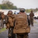 The image shows a group of military personnel, likely U.S. Marines based on their woodland MARPAT camouflage uniforms, assembled on a paved area such as a tarmac or road. The central figure, whose backpack bears the name tape 'LINCOLN,' is wearing a cover (patrol cap) and carrying a large MOLLE-style tactical pack. Personnel are lined up on both sides in the background, suggesting a formation, inspection, or pre-deployment staging event.