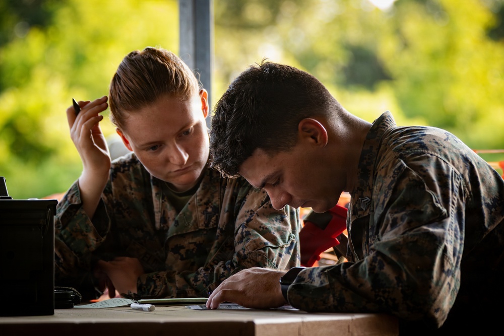 Two military personnel in camouflage uniforms are working together at a table, reviewing documents or paperwork. Both individuals are wearing what appears to be MARPAT (Marine Pattern) digital camouflage uniforms, consistent with U.S. Marine Corps attire. The setting appears to be an outdoor or semi-outdoor workspace with green foliage visible in the background.