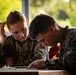 Two military personnel in camouflage uniforms are working together at a table, reviewing documents or paperwork. Both individuals are wearing what appears to be MARPAT (Marine Pattern) digital camouflage uniforms, consistent with U.S. Marine Corps attire. The setting appears to be an outdoor or semi-outdoor workspace with green foliage visible in the background.