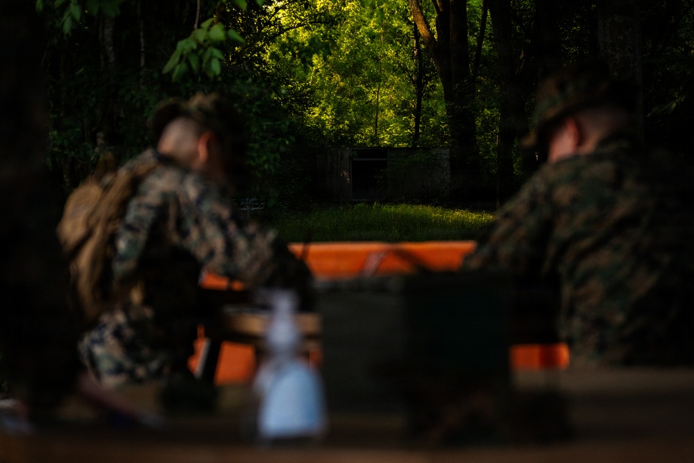 Two individuals wearing military camouflage uniforms are seated at what appears to be a wooden table or bench in a wooded outdoor setting. The image is intentionally shallow in depth of field, with the foreground and background subjects out of focus. Green military-style containers and other equipment are visible on the table surface.