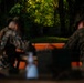 Two individuals wearing military camouflage uniforms are seated at what appears to be a wooden table or bench in a wooded outdoor setting. The image is intentionally shallow in depth of field, with the foreground and background subjects out of focus. Green military-style containers and other equipment are visible on the table surface.