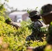 The image shows military personnel in camouflage uniforms concealed within dense green vegetation, appearing to conduct a field training exercise or tactical movement drill. At least three individuals are visible, wearing digital camouflage pattern uniforms and boonie hats with natural foliage attached for additional concealment. A structure and vehicle are visible in the blurred background, suggesting a semi-urban or base training environment.
