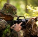 A soldier in digital camouflage uniform is positioned behind a fallen log in a forested environment, aiming a rifle forward. The individual has vegetation tucked into their hat and uniform as natural camouflage concealment. The scene appears to depict a military field training exercise or tactical operation in a woodland setting.