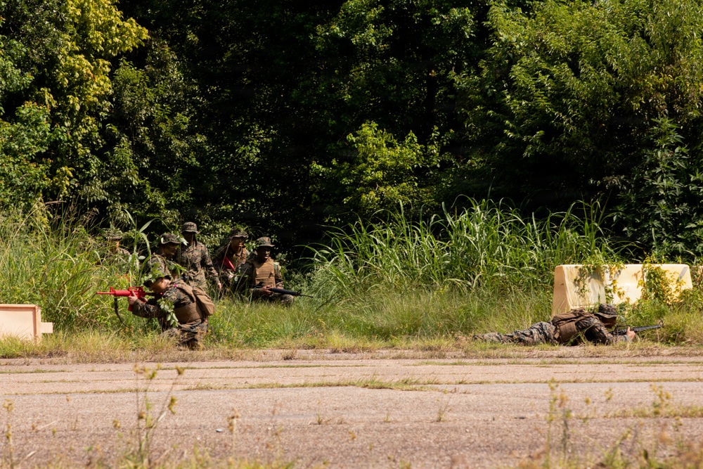 The image shows a group of military personnel in camouflage uniforms conducting what appears to be a tactical training exercise in an outdoor environment. Several soldiers are positioned in a crouching or prone formation along a grassy area adjacent to a paved surface, with at least one individual lying flat in a firing position. Training weapons with red markings (indicating simunition or MILES gear) are visible, confirming this is a live-force-on-force training scenario.