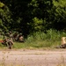 The image shows a group of military personnel in camouflage uniforms conducting what appears to be a tactical training exercise in an outdoor environment. Several soldiers are positioned in a crouching or prone formation along a grassy area adjacent to a paved surface, with at least one individual lying flat in a firing position. Training weapons with red markings (indicating simunition or MILES gear) are visible, confirming this is a live-force-on-force training scenario.