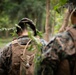 The image shows multiple military personnel in woodland camouflage uniforms moving through a forested environment. The central figure has natural foliage tucked into their helmet as additional camouflage. The scene appears to depict a field training exercise or patrol in a wooded area.