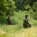 Two individuals in military camouflage uniforms are kneeling in a grassy clearing surrounded by dense green vegetation. One person faces the camera and appears to be communicating using hand signals or gestures, while the other faces away. The setting and attire suggest a military training exercise or field operation.