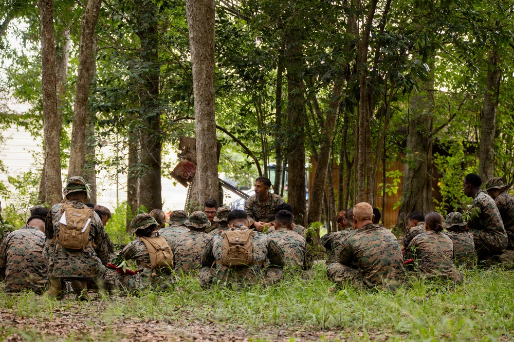 A group of military personnel in camouflage uniforms are gathered in a wooded outdoor area, seated on the ground in a semicircle formation. One individual appears to be standing or positioned facing the group, suggesting a briefing, instruction, or after-action review is taking place. The setting appears to be a training environment with trees providing shade and cover.