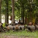 A group of military personnel in camouflage uniforms are gathered in a wooded outdoor area, seated on the ground in a semicircle formation. One individual appears to be standing or positioned facing the group, suggesting a briefing, instruction, or after-action review is taking place. The setting appears to be a training environment with trees providing shade and cover.