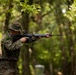 A soldier in digital camouflage uniform is aiming a rifle in a wooded training environment. The individual is wearing MARPAT (Marine pattern) camouflage with natural vegetation attached for concealment, suggesting a field training exercise. Additional personnel in similar uniforms are partially visible in the background.