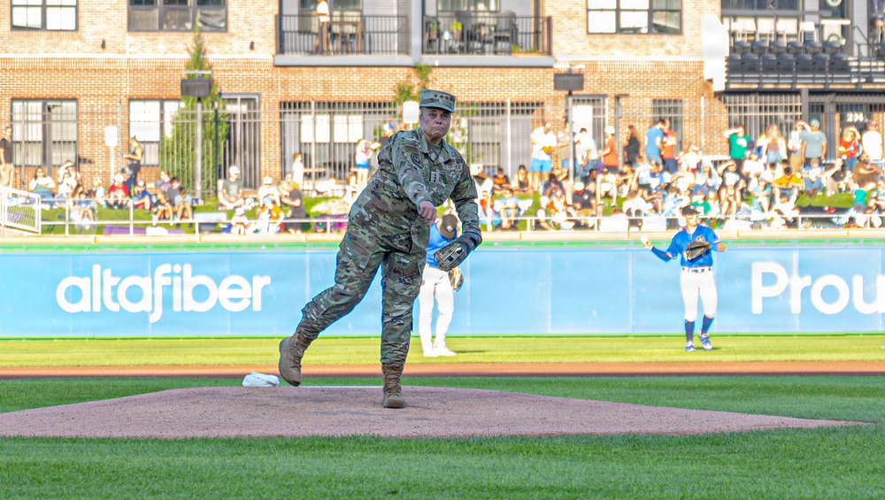A U.S. military service member in full camouflage (OCP/MultiCam) uniform is throwing a ceremonial first pitch at a minor league baseball stadium. The event appears to be a military appreciation night, with spectators filling the stands behind an outfield wall featuring an 'altafiber' advertisement. A baseball player in a blue uniform stands in the background near the outfield.