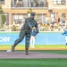 A U.S. military service member in full camouflage (OCP/MultiCam) uniform is throwing a ceremonial first pitch at a minor league baseball stadium. The event appears to be a military appreciation night, with spectators filling the stands behind an outfield wall featuring an 'altafiber' advertisement. A baseball player in a blue uniform stands in the background near the outfield.