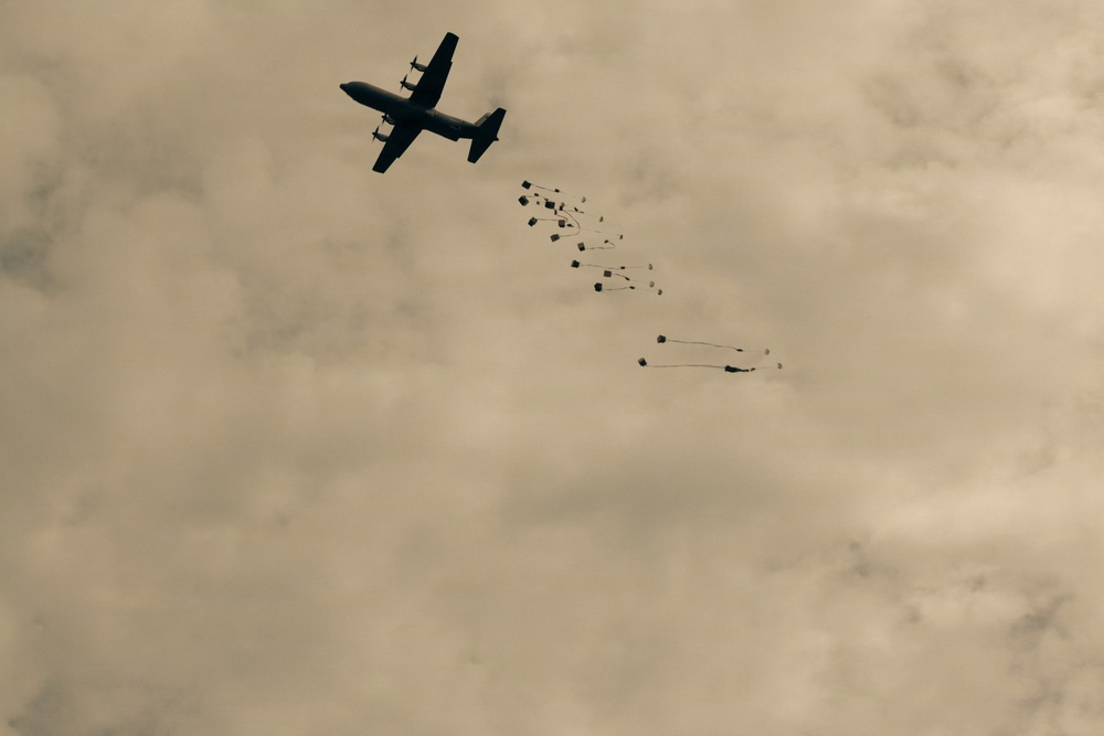 A military cargo aircraft, consistent with a C-130 Hercules silhouette, is captured mid-flight performing an aerial delivery operation. Multiple cargo bundles equipped with small extraction or drogue parachutes are visible trailing behind and below the aircraft. The image has a sepia/overcast tone with a cloudy sky background.