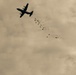 A military cargo aircraft, consistent with a C-130 Hercules silhouette, is captured mid-flight performing an aerial delivery operation. Multiple cargo bundles equipped with small extraction or drogue parachutes are visible trailing behind and below the aircraft. The image has a sepia/overcast tone with a cloudy sky background.