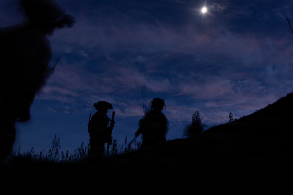 A nighttime silhouette photograph showing what appear to be armed military or tactical personnel standing on elevated terrain under a moonlit sky. The figures are rendered entirely in dark silhouette against a deep blue twilight sky with scattered clouds. A bright moon or light source is visible in the upper right portion of the frame.
