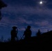 A nighttime silhouette photograph showing what appear to be armed military or tactical personnel standing on elevated terrain under a moonlit sky. The figures are rendered entirely in dark silhouette against a deep blue twilight sky with scattered clouds. A bright moon or light source is visible in the upper right portion of the frame.
