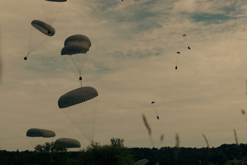 The image shows a large-scale military airborne operation with numerous parachutes deployed across the sky. Multiple round, olive-drab/gray military parachutes are visible at various altitudes, each carrying suspended loads or personnel. The scene is set against an overcast sky with a treeline silhouetted along the horizon.