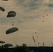 The image shows a large-scale military airborne operation with numerous parachutes deployed across the sky. Multiple round, olive-drab/gray military parachutes are visible at various altitudes, each carrying suspended loads or personnel. The scene is set against an overcast sky with a treeline silhouetted along the horizon.