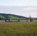 A group of military personnel in camouflage uniforms are conducting a field exercise across an open grassy plain, with purple/violet colored smoke deployed in the background. The soldiers appear to be advancing or maneuvering across the terrain in a tactical formation. The scene is set against a backdrop of green rolling hills and a partly cloudy sky.