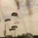 A military airdrop operation is captured mid-execution, showing multiple olive-drab parachutes descending with cargo loads suspended beneath them. Several parachutes are still airborne at various altitudes while at least one has already landed on the ground to the right, with its canopy still inflated. The scene takes place over an open grassy field bordered by trees under an overcast sky.