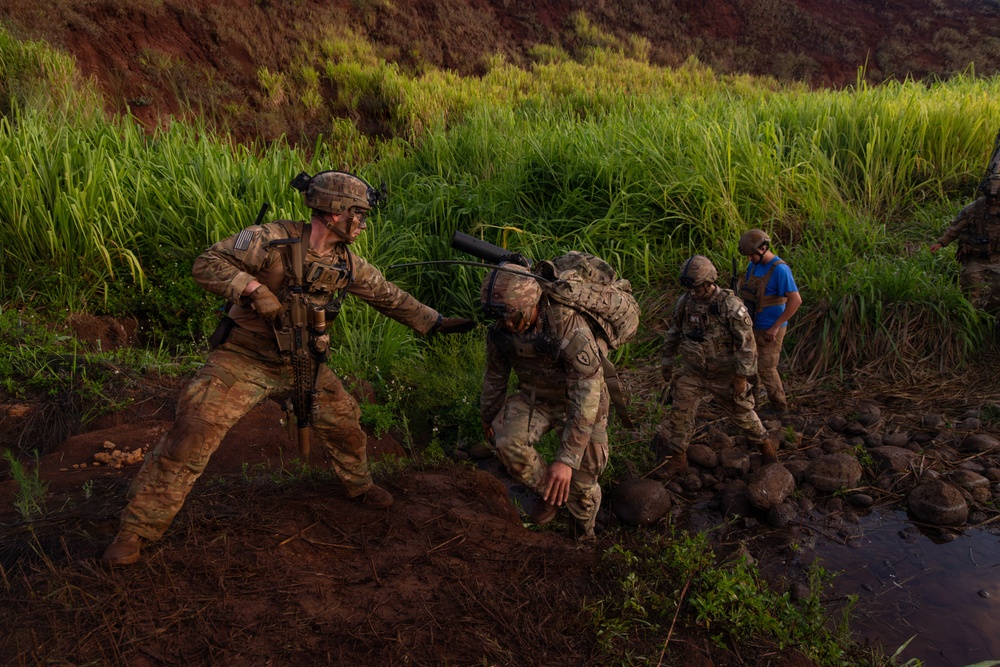 Military personnel in full combat gear navigate difficult terrain near a muddy stream or water crossing in a lush, tropical environment. One soldier is assisting or steadying another who is bent forward under the weight of a heavy pack, while additional personnel are visible in the background. The setting appears to be a training exercise given the controlled nature of the movement and presence of a civilian in a blue shirt.