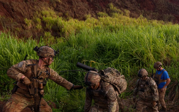 Military personnel in full combat gear navigate difficult terrain near a muddy stream or water crossing in a lush, tropical environment. One soldier is assisting or steadying another who is bent forward under the weight of a heavy pack, while additional personnel are visible in the background. The setting appears to be a training exercise given the controlled nature of the movement and presence of a civilian in a blue shirt.