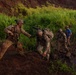 Military personnel in full combat gear navigate difficult terrain near a muddy stream or water crossing in a lush, tropical environment. One soldier is assisting or steadying another who is bent forward under the weight of a heavy pack, while additional personnel are visible in the background. The setting appears to be a training exercise given the controlled nature of the movement and presence of a civilian in a blue shirt.