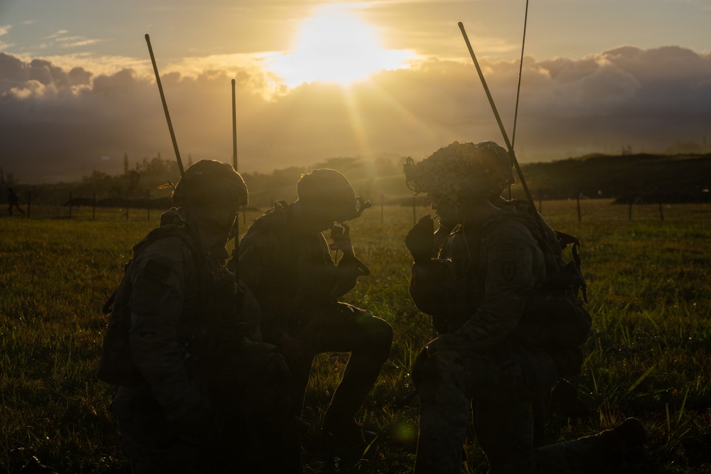 Three military personnel in full combat gear are silhouetted against a dramatic sunrise or sunset in an open grassy field. The figures appear to be in a tactical huddle or briefing, kneeling and crouching together. The backlighting creates a strong silhouette effect, obscuring individual facial features and fine equipment details.