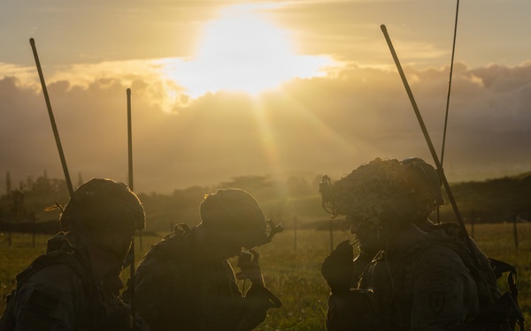 Three military personnel in full combat gear are silhouetted against a dramatic sunrise or sunset in an open grassy field. The figures appear to be in a tactical huddle or briefing, kneeling and crouching together. The backlighting creates a strong silhouette effect, obscuring individual facial features and fine equipment details.