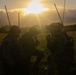 Three military personnel in full combat gear are silhouetted against a dramatic sunrise or sunset in an open grassy field. The figures appear to be in a tactical huddle or briefing, kneeling and crouching together. The backlighting creates a strong silhouette effect, obscuring individual facial features and fine equipment details.