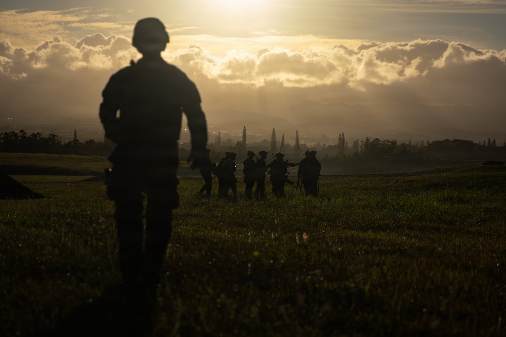 A dramatic silhouette photograph showing military personnel in a field during golden hour or sunrise/sunset conditions. One soldier stands in the foreground out of focus, while a group of approximately 5-7 armed soldiers are gathered in the middle distance. The scene is set against a luminous sky with dramatic cloud formations and a treeline on the horizon.