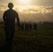 A dramatic silhouette photograph showing military personnel in a field during golden hour or sunrise/sunset conditions. One soldier stands in the foreground out of focus, while a group of approximately 5-7 armed soldiers are gathered in the middle distance. The scene is set against a luminous sky with dramatic cloud formations and a treeline on the horizon.
