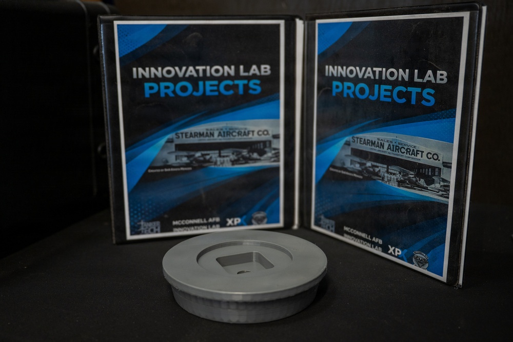 The image shows two open binders labeled 'Innovation Lab Projects' standing upright on a dark surface, both featuring a historical photograph of Stearman Aircraft Co. and branding for McConnell AFB Innovation Lab and XP. In front of the binders sits a small metallic disc-shaped object, likely a machined or 3D-printed component. The overall composition suggests a display or presentation of innovation lab work products.