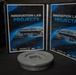 The image shows two open binders labeled 'Innovation Lab Projects' standing upright on a dark surface, both featuring a historical photograph of Stearman Aircraft Co. and branding for McConnell AFB Innovation Lab and XP. In front of the binders sits a small metallic disc-shaped object, likely a machined or 3D-printed component. The overall composition suggests a display or presentation of innovation lab work products.