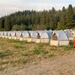 A large field contains dozens of uniformly arranged dome-style tents in tan and brown colors, set up in organized rows. The setting appears to be a temporary base camp, likely for wildfire firefighters or emergency personnel, given the systematic layout and forest backdrop. An orange traffic cone is visible among the tents, suggesting organized logistics and site management.