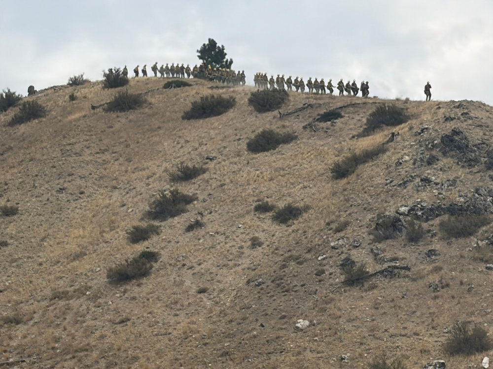 A large group of individuals wearing yellow/tan protective gear and hard hats are lined up along the ridgeline of a dry, arid hillside. The terrain shows signs of recent burning, with blackened patches visible on the rocky slope. The formation and attire are consistent with a wildland firefighting crew conducting operations on or near a fire line.