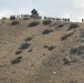 A large group of individuals wearing yellow/tan protective gear and hard hats are lined up along the ridgeline of a dry, arid hillside. The terrain shows signs of recent burning, with blackened patches visible on the rocky slope. The formation and attire are consistent with a wildland firefighting crew conducting operations on or near a fire line.
