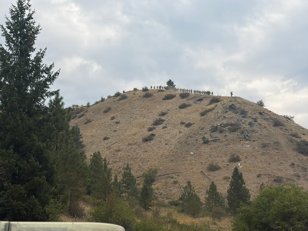 A large group of people is visible along the ridgeline of a dry, rocky hill, appearing to move in a single-file line or column formation. The hill is sparsely vegetated with scrub brush and a few trees, set against an overcast sky. The scene is photographed from a distance, making individual figures small but collectively visible as a procession along the summit.