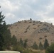 A large group of people is visible along the ridgeline of a dry, rocky hill, appearing to move in a single-file line or column formation. The hill is sparsely vegetated with scrub brush and a few trees, set against an overcast sky. The scene is photographed from a distance, making individual figures small but collectively visible as a procession along the summit.